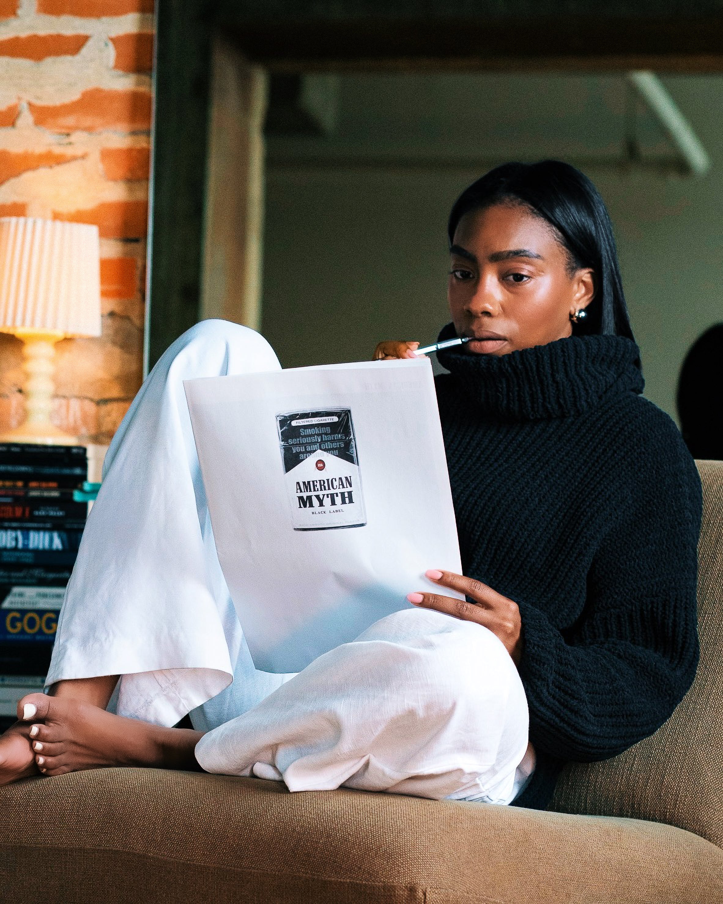 Woman sitting on a couch holding a magazine titled 'American Myth' in a cozy indoor setting.
